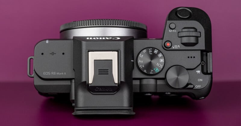 A top-down view of a black mirrorless camera body labeled "EOS R8 Mark II" with a mode dial, shutter button, and hot shoe against a solid purple background.