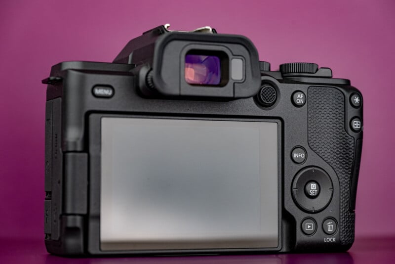 The back of a black digital mirrorless camera with a large LCD screen, viewfinder, and various control buttons against a solid magenta background.