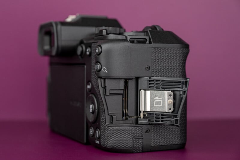 A black digital camera is shown from the rear-right angle with its memory card slot door open, revealing the internal compartment.