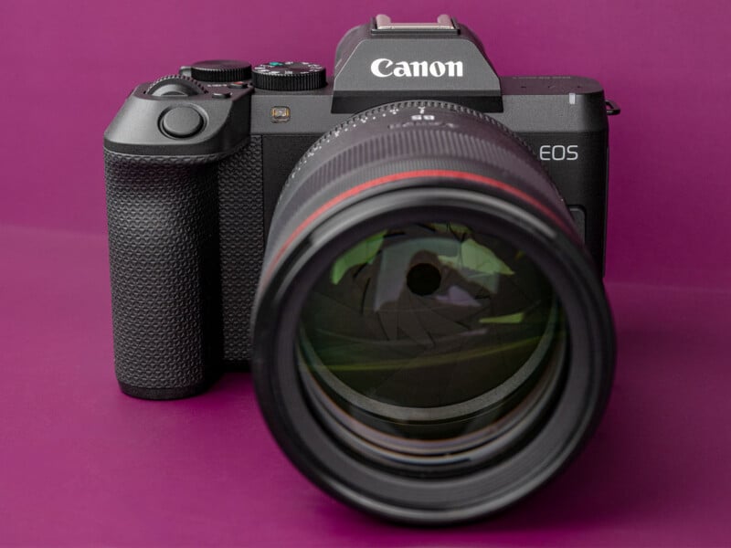 A black Canon EOS mirrorless camera with a large lens attached, set against a solid purple background.