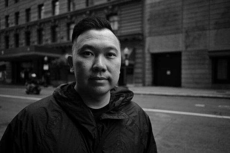 A man with short, dark hair and a serious expression stands outdoors in front of a city building, wearing a dark hooded jacket.