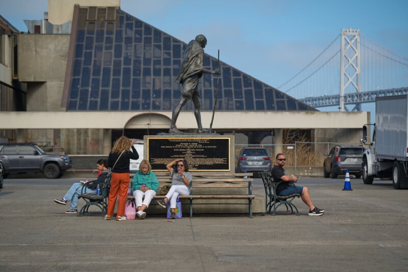A bronze statue of Mahatma Gandhi stands on a pedestal behind a park bench where four people are sitting and one is standing, with the San Francisco-Oakland Bay Bridge visible in the background.
