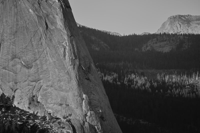 A large, steep granite rock face rises on the left, overlooking a dense forest valley under a clear sky.