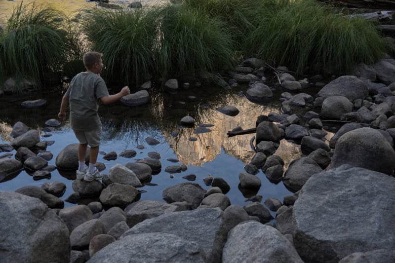 A young boy in a t-shirt and shorts stands on a rock in a shallow stream, looking at the reflection of a mountain in the water.