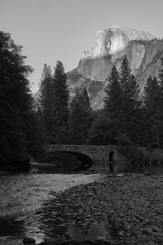 A stone arch bridge spans a river in front of a large, steep mountain peak, framed by tall pine trees.