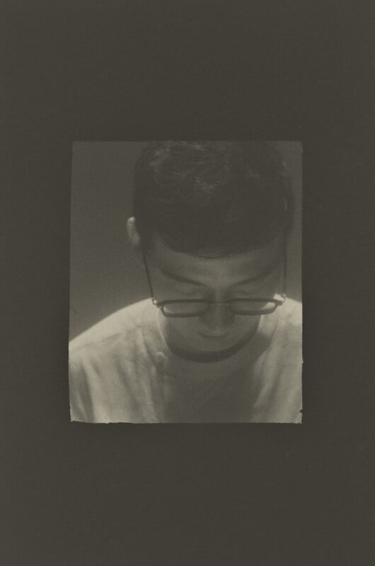 A person with short dark hair wearing glasses looks downward in a sepia-toned portrait.