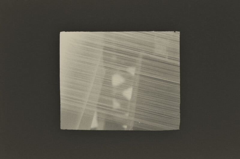 A rectangular, sepia-toned print with a textured, streaked appearance features faint, geometric shapes layered behind diagonal lines.