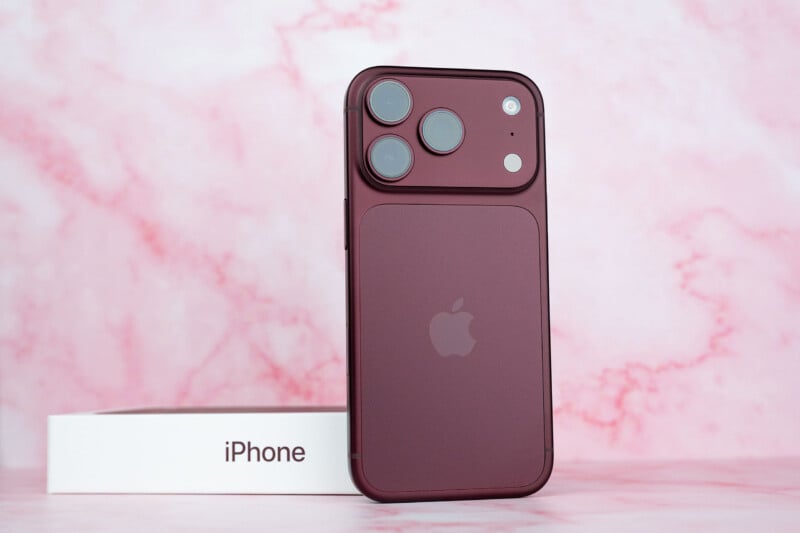 A dark red smartphone stands upright in front of its white box on a pink marble-patterned surface.