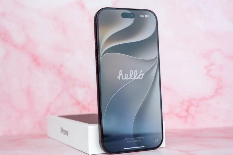 A dark-colored smartphone stands upright on its white retail box against a pink marble-patterned background, displaying a "hello" welcome screen in Hungarian.