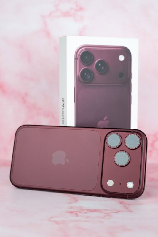 A dark red smartphone rests horizontally in front of its matching product box on a pink marble-patterned surface.