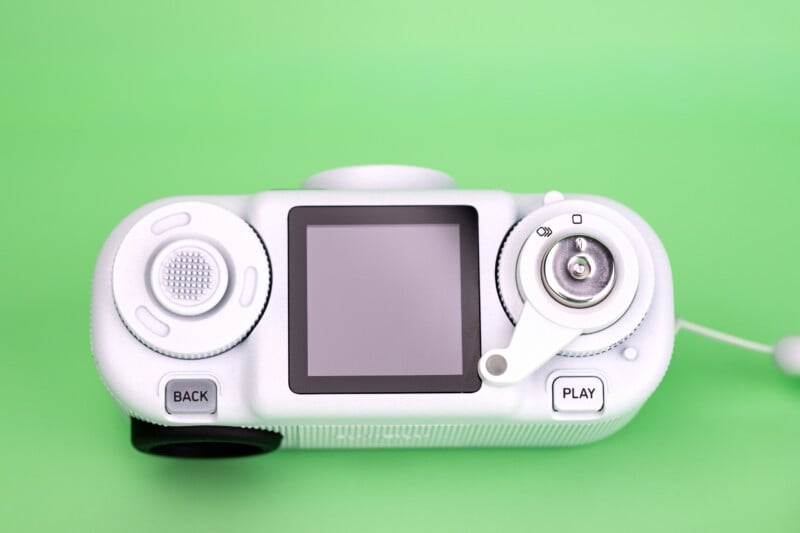 A white digital camera with a square screen, a circular dial, a crank-style lever, and buttons labeled "BACK" and "PLAY" sits against a solid green background.