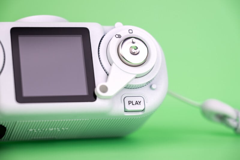 A close-up view of the back of a white digital camera against a solid green background, showing the screen, a play button, and a circular control dial.