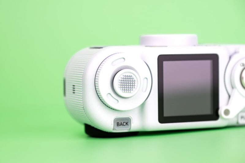 A white digital camera sits on a solid light green background, showing its rear screen, a textured control dial, and a button labeled "BACK.