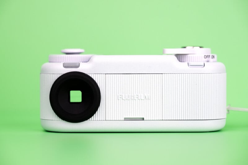 A white Fujifilm camera sits against a solid light green background, showing its viewfinder, a textured body, and an on/off switch.