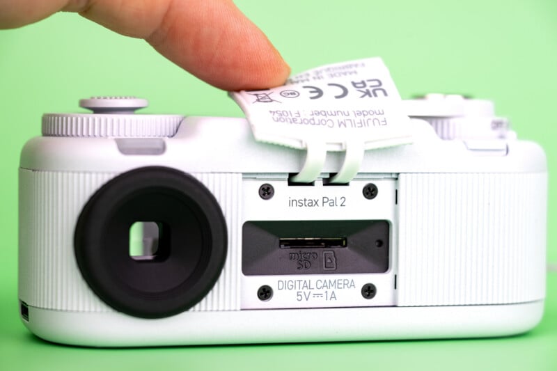 A finger lifts a rubber flap on the back of a white Instax Pal 2 digital camera, revealing a microSD card slot.