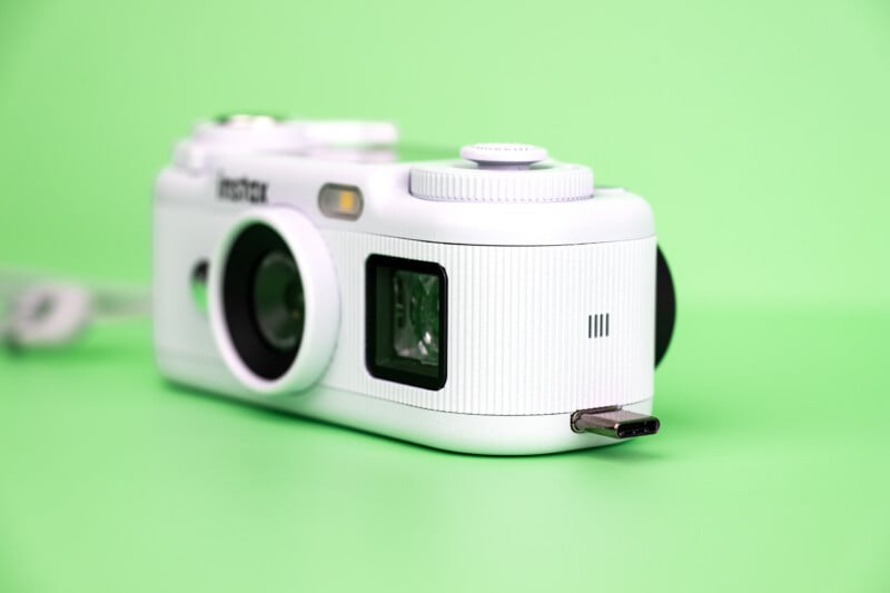 A white Instax digital camera sits on a solid light green surface with a USB-C connector protruding from its side.