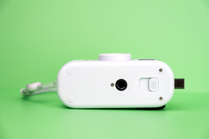 The bottom view of a white, oval-shaped digital camera sits on a green surface, showing a tripod mount and a sliding latch.