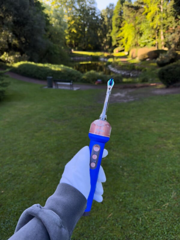 A person wearing a white glove and grey sleeve holds a blue electric toothbrush against a background of a park with a pond and trees.