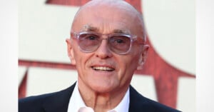 A bald, older man with light skin and thin eyebrows smiles while wearing clear-framed glasses, a white collared shirt, and a black blazer.