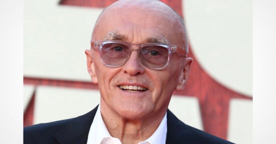 A bald, older man with light skin and thin eyebrows smiles while wearing clear-framed glasses, a white collared shirt, and a black blazer.