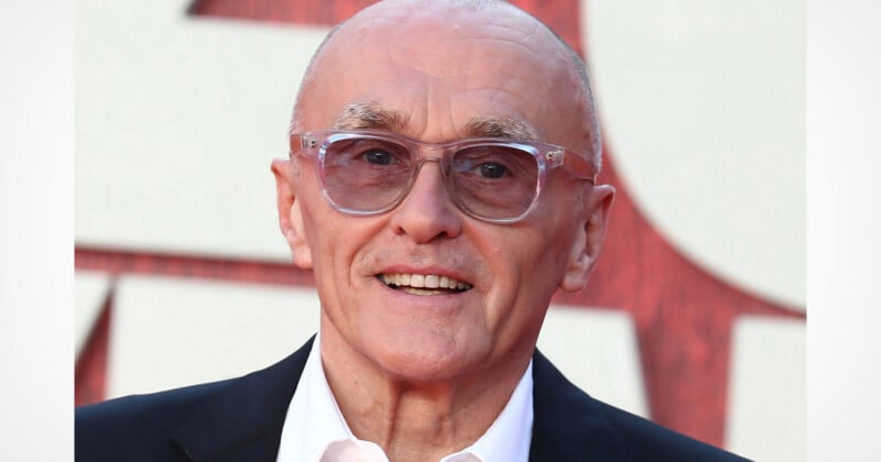 A bald, older man with light skin and thin eyebrows smiles while wearing clear-framed glasses, a white collared shirt, and a black blazer.