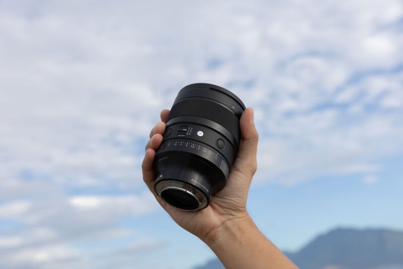 A hand holds a black camera lens against a background of a cloudy sky and distant mountains.