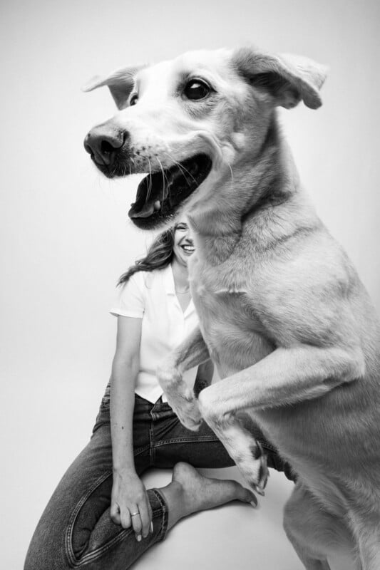 A black-and-white portrait of a woman sitting on the floor while a large dog stands in the foreground, partially obscuring her face.