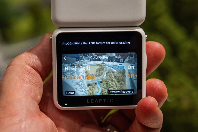 A hand holds a small, white digital camera displaying a menu screen with options for HLG(10bit) and P-LOG(10bit) color settings.