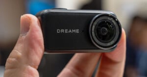A hand holds a small, rectangular black camera with the word "DREAME" printed on the front next to a circular lens.