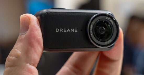 A hand holds a small, rectangular black camera with the word "DREAME" printed on the front next to a circular lens.