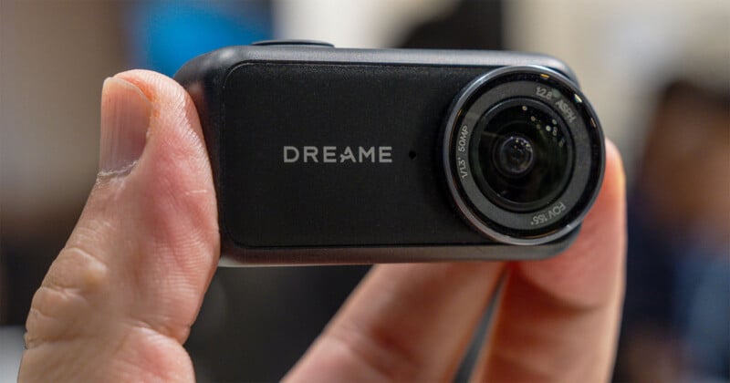 A hand holds a small, rectangular black camera with the word "DREAME" printed on the front next to a circular lens.