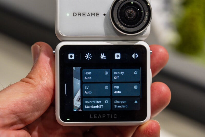 A hand holds a white, square-shaped camera with a digital screen displaying a settings menu featuring options for HDR, Beauty, EV, WB, Color/Filter, and Sharpen.