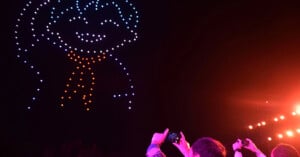 A drone light show in the night sky forms the outline of a smiling character wearing a scarf, while spectators in the foreground capture the display with their smartphones.