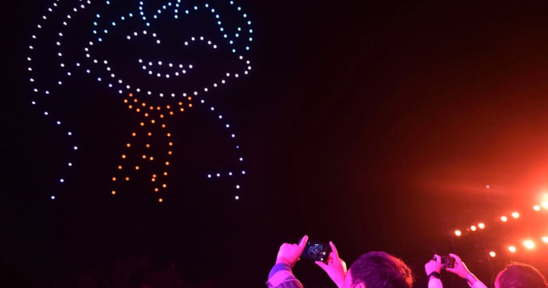 A drone light show in the night sky forms the outline of a smiling character wearing a scarf, while spectators in the foreground capture the display with their smartphones.