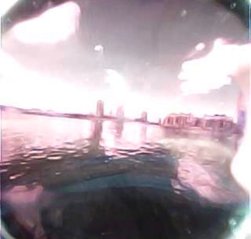 A distorted, fisheye-lens view shows a body of water with a bridge and buildings visible in the distance under a bright sky.