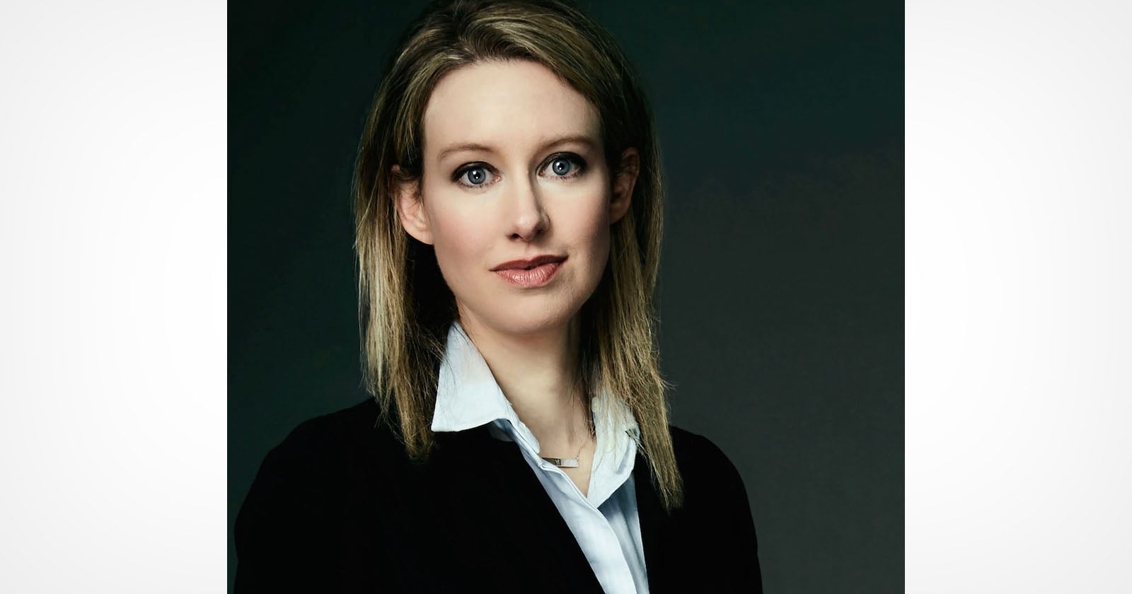 A portrait of Elizabeth Holmes with blonde hair, wearing a white collared shirt and a black blazer against a dark background.