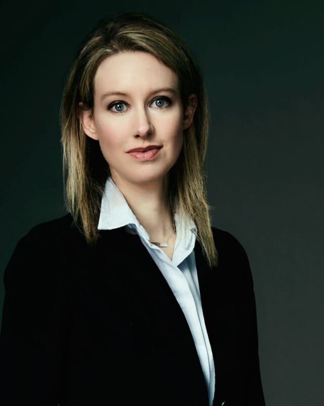 A woman with shoulder-length blonde hair and blue eyes, wearing a white collared shirt under a black blazer, looks directly at the camera against a dark gray background.