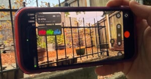 A hand holds a smartphone displaying a professional camera app interface with a live waveform monitor, recording settings, and a view of a backyard with a metal fence and autumn leaves.