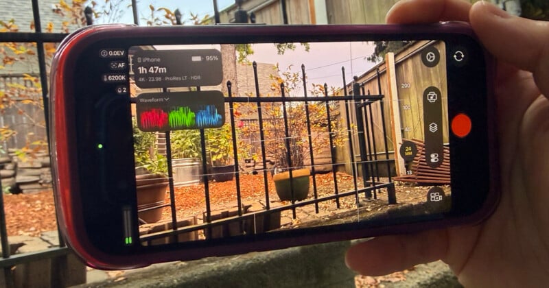A hand holds a smartphone displaying a professional camera app interface with a live waveform monitor, recording settings, and a view of a backyard with a metal fence and autumn leaves.
