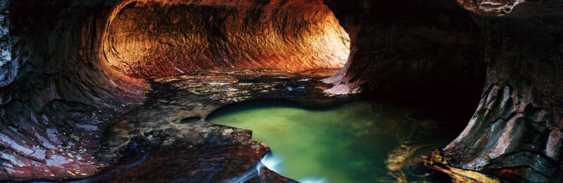 A smooth, carved sandstone canyon features a glowing orange interior and a calm, turquoise pool of water in the foreground.