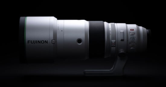A silver Fujinon telephoto lens is positioned horizontally against a black background, featuring a tripod mount and various control switches on its side.