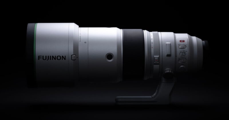 A silver Fujinon telephoto lens is positioned horizontally against a black background, featuring a tripod mount and various control switches on its side.