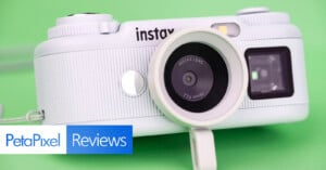 A white Instax digital camera sits on a solid green background, with a PetaPixel Reviews logo in the bottom left corner.