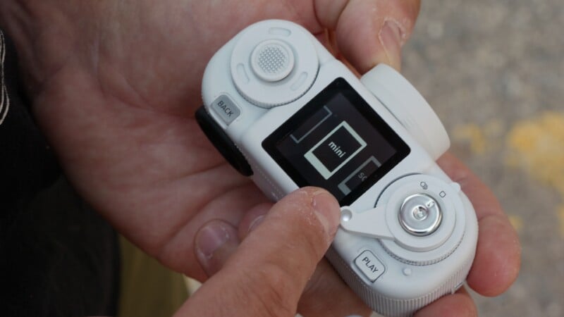 A person holds a white, handheld electronic device with a small screen displaying the word "mini" and a "PLAY" button.
