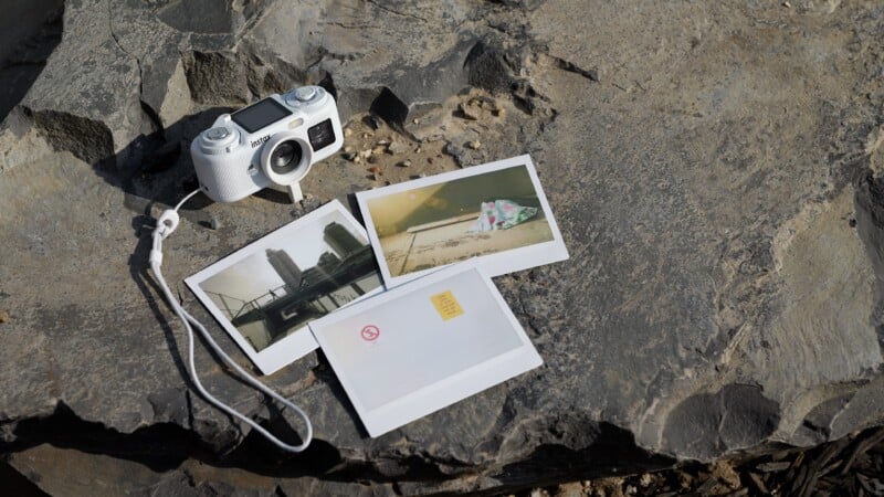 A white digital instant camera with a wrist strap sits on a rocky surface next to three printed photographs.
