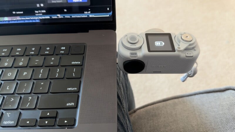 A light gray Fujifilm digital camera is plugged directly into the side of a laptop, displaying a full battery icon on its screen.