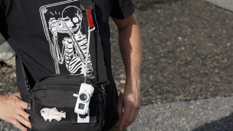 A person wearing a black t-shirt with a skeleton graphic and a black crossbody bag with a small white camera attached to the strap.