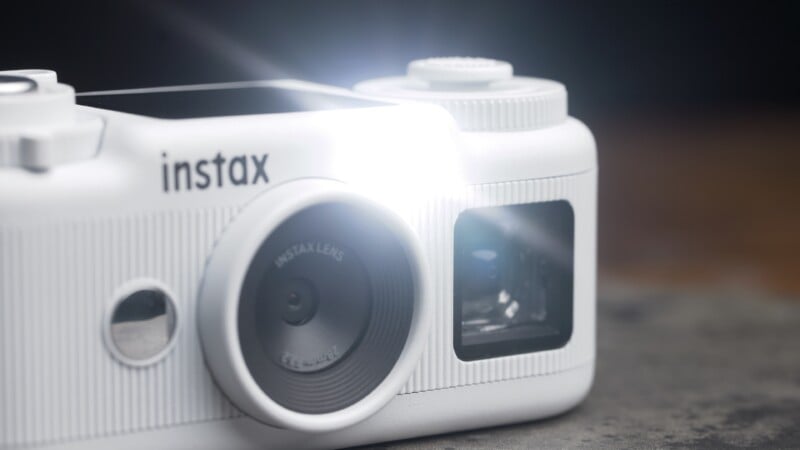 A close-up, angled view of a white Instax digital camera with a bright flash emitting from the front, showing the lens and a small mirror.