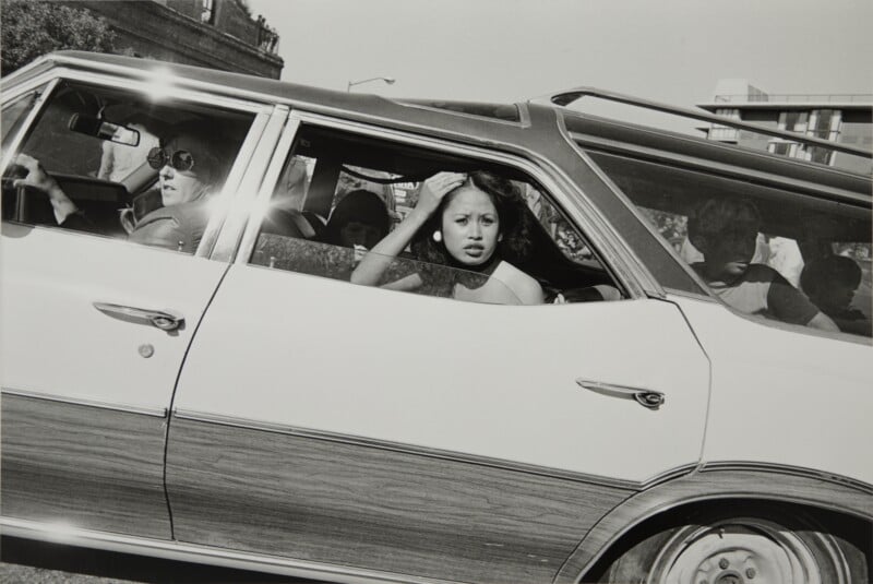 A black-and-white photograph shows a woman with dark hair looking out the window of a wood-paneled station wagon, with a driver wearing sunglasses and other passengers visible inside.