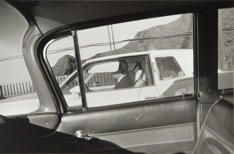 A view from inside a car looking out the window at a white car driving alongside, with a person visible in the driver's seat.